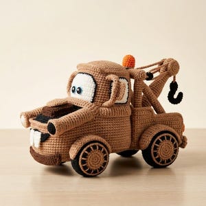 May include: A brown crocheted tow truck toy featuring large white eyes with blue pupils, a black front bumper, and two white teeth. The vehicle includes a rear crane arm with a black hook and a small orange light on top, all crafted with yarn.
