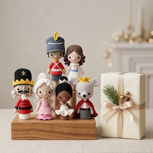 Puede incluir: Una colección de seis muñecos de ganchillo que representan personajes de El Cascanueces, incluyendo un soldado, un rey ratón y bailarines, dispuestos sobre un soporte de madera rectangular junto a un regalo color crema con cinta de raso.