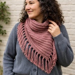 May include: A dusty rose triangular scarf featuring a ribbed knit texture and long fringe trim along the bottom edges. The scarf is draped around the neck, showcasing the soft yarn construction and the consistent pattern of the vertical ribbing.