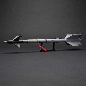 May include: A scale model of a grey missile featuring a black nose cone, triangular fins, and a cylindrical body with various markings. A small red tag with white text hangs from the center, and the model is supported by a black stand.