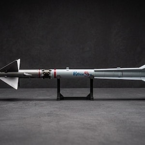 Könnte beinhalten: Ein graues Python 5 Raketenmodell mit gelber Nasenkegelspitze, weißen Finnen und einem roten Anhänger mit der Aufschrift REMOVE BEFORE FLIGHT. Der zylindrische Körper zeigt den Schriftzug Python 5 in Blau und Rot auf einem schwarzen Ständer.