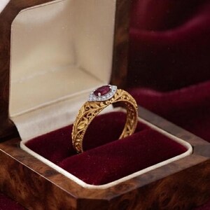Può includere: Un anello in oro con una fascia dal motivo a foglie intrecciate e una gemma rossa centrale taglio marquise, circondata da un alone di piccole pietre bianche. L'anello è presentato in un cofanetto di legno con interno in velluto rosso.