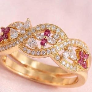 May include: A gold ring featuring an intertwined band design encrusted with small clear stones. The ring is accented with marquise-cut clear stones and small, four-petaled flower motifs composed of deep pink, marquise-cut gemstones.