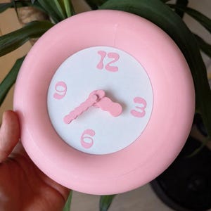 May include: A round wall clock with a thick, matte pink frame and a white circular face. The clock features pink, wavy-edged hands and large, stylized pink numerals for 12, 3, 6, and 9 positioned at the cardinal points on the white background.