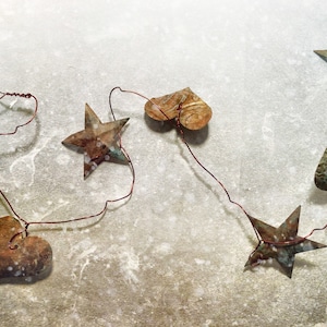May include: A rustic garland featuring metal stars and hearts strung on a thin wire. The metal has a weathered, aged appearance with shades of brown, green, and white. The garland is designed for decorative purposes.