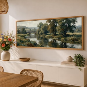 May include: A panoramic landscape painting in a light wood frame depicting a river flowing under a stone bridge, surrounded by lush green trees and rolling hills. The artwork features muted earth tones, soft lighting, and reflections on the water.