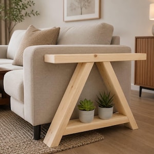 May include: A light-coloured wooden side table with an A-frame design, featuring a flat top surface and a lower shelf that holds two small potted succulent plants. The table is positioned directly against the side of a beige upholstered sofa.