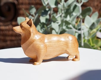 May include: A wooden carving of a corgi dog, standing on a white surface. The carving is made of light brown wood and has a smooth finish.
