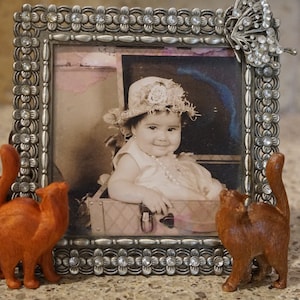 May include: A silver picture frame with a rhinestone border and a butterfly design. The frame holds a black and white photo of a young girl wearing a hat and sitting in a suitcase. Three wooden cat figurines are placed around the frame.
