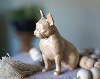 Può includere: Una statuetta in legno di colore chiaro intagliata a forma di bulldog francese. La scultura presenta venature del legno visibili, orecchie a punta, un muso corto e una superficie liscia e lucida, che cattura la postura tozza della razza.