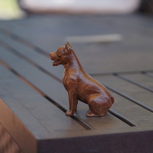 May include: A wooden figurine of a dog sitting on a dark brown wooden surface. The dog is brown and has a short, smooth coat. The figurine is detailed and realistic.
