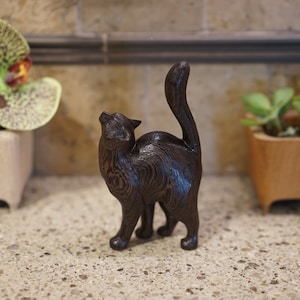 Könnte beinhalten: Eine dunkelbraune Holzkatzenfigur mit hoch erhobenem Schwanz, die auf einer hellbraunen, gesprenkelten Oberfläche steht.