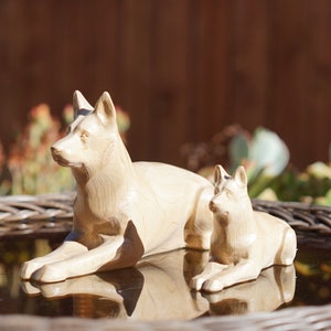 Könnte beinhalten: Zwei Holzskulpturen von Hunden, eine Mutter und ein Welpe, werden auf einer reflektierenden Oberfläche gezeigt. Der größere Hund liegt, während der kleinere daneben liegt. Die Skulpturen sind hell und detailliert, mit natürlicher Holzmaserung.