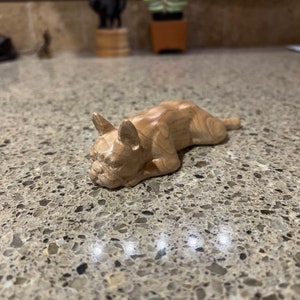 May include: A wooden carving of a French bulldog lying down. The carving is made of light brown wood with dark brown wood grain.