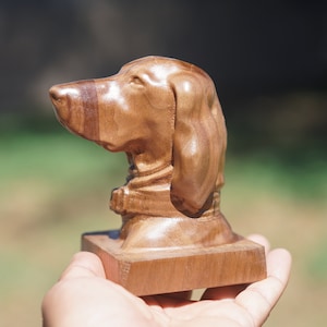 May include: A wooden sculpture of a dog's head, carved from light brown wood with darker brown stripes. The sculpture is on a small wooden base and is being held in someone's hand.