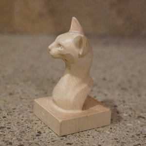 May include: A wooden sculpture of a cat's head and neck, carved in a simple style. The sculpture is painted in a light wood colour and is sitting on a small wooden base.