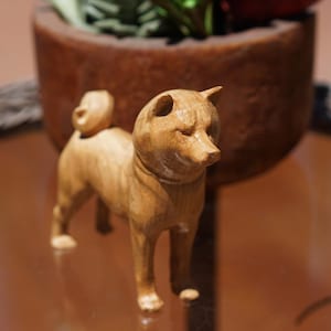 May include: A wooden figurine of a Shiba Inu dog, standing on a reflective surface. The dog is carved from light-colored wood, with detailed features. The background includes a brown pot with greenery.