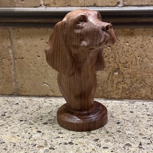 May include: A detailed wooden sculpture of a dog's head, showcasing the breed's features. The sculpture is a rich brown color and is mounted on a round base. The dog's head is turned slightly to the side, with its ears hanging down.