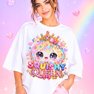 Może przedstawiać: Biały t-shirt z kolorową grafiką postaci z kreskówki o dużych, błyszczących oczach, noszącej złotą koronę i różową kokardę. Poniżej znajduje się napis SQUISHY QUEEN wykonany wielokolorową czcionką przypominającą bąbelki.