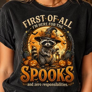 May include: A black t-shirt featuring a graphic of a raccoon wearing a witch hat and holding a lantern. The design includes pumpkins, bats, a full moon, and the text: FIRST OF ALL I'M HERE FOR THE SPOOKS and zero responsibilities.