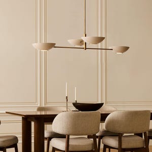 Puede incluir: Una lámpara de araña moderna con una varilla horizontal de latón delgada suspendida de un vástago vertical central, que sostiene cuatro pantallas blancas mate en forma de cuenco que emiten una luz suave hacia abajo, creando un diseño lineal.
