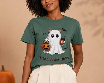 May include: A dark green short-sleeve t-shirt featuring a graphic of a cat dressed as a ghost, holding a jack-o'-lantern bucket and a coffee cup, surrounded by small bats. The text "Treats Before Tricks" is printed in a dark red font below.