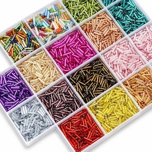 Puede incluir: Una bandeja organizadora de plástico blanco llena de un surtido de pequeñas cuentas de vidrio cilíndricas en varios colores, incluyendo oro metálico, plata, cobre, negro, rojo, amarillo, rosa, morado, verde, azul y multicolor iridiscente.