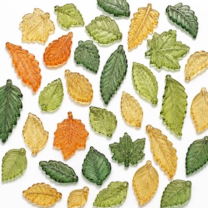 May include: A collection of translucent glass leaf beads in various shapes and sizes, featuring shades of amber, orange, light green, and dark green. Each bead has a textured surface with detailed vein patterns and a small hole at the base.
