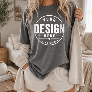 Może przedstawiać: Ciemnoszary t-shirt z krótkim rękawem z białą okrągłą grafiką na klatce piersiowej z napisem YOUR DESIGN HERE oraz NLH STUDIOS oddzielonymi małym sercem. Koszulka ma okrągły dekolt i luźny krój, zestawiona z jasnobeżowymi szortami w komplecie.