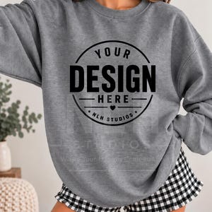 May include: A heather grey crewneck sweatshirt featuring a black circular graphic on the chest with the text YOUR DESIGN HERE and NLH STUDIOS with a small heart icon. The sweatshirt is paired with black and white gingham print ruffled shorts.