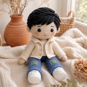 May include: A handmade crochet doll featuring a person with black yarn hair, rosy cheeks, and a neutral expression. The doll wears a cream-colored button-up cardigan over a white shirt, blue pants with green patches on the knees, and white shoes.
