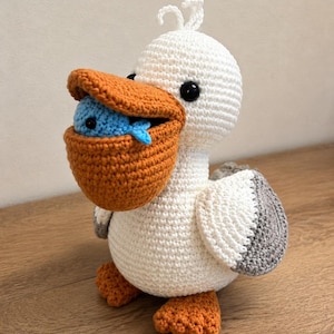 May include: A crocheted pelican toy with a white body, orange feet, and a large orange beak holding a small blue fish. The pelican has black bead eyes, a small tuft of white yarn on its head, and grey and white wings folded at its sides.