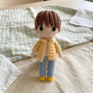 May include: A handmade crochet doll featuring brown yarn hair, black bead eyes, and pink cheeks. The doll wears a yellow button-down shirt, light blue trousers, and yellow shoes. The figure is crafted with detailed yarn stitching throughout.