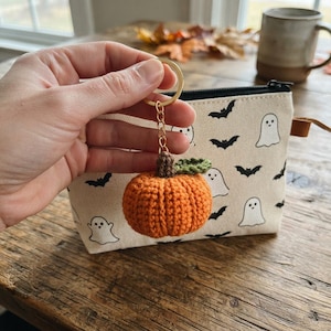 May include: A crocheted orange pumpkin keychain with a brown stem and a green leaf attached to a gold-toned metal ring. Behind the keychain is a cream-colored zippered pouch featuring a repeating pattern of black bats and white ghosts.