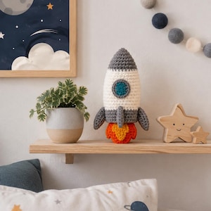 Pode incluir: Um brinquedo de foguete em crochê com topo pontiagudo cinza, corpo cilíndrico branco com uma janela circular azul, barbatanas cinzas e uma base de chamas laranjas e amarelas. O foguete está posicionado em uma prateleira de madeira.