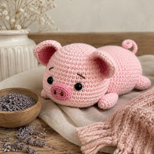 May include: A light pink crocheted pig toy with a rounded body, large ears, a snout with two dark nostrils, and black bead eyes. The pig has a small curled tail and is crafted with a textured yarn stitch, featuring embroidered eyebrows above the eyes.
