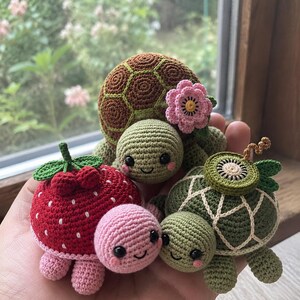 Puede incluir: Tres pequeñas figuras de tortugas tejidas a ganchillo. Una tiene un caparazón marrón con una flor rosa, otra es roja con puntos blancos como una fresa, y otra es verde con una rodaja de kiwi en el caparazón. Tienen caras sonrientes y ojos negros.