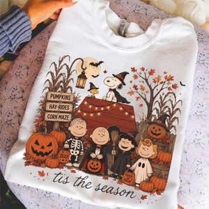Puede incluir: Una sudadera blanca de cuello redondo con un gráfico colorido de Halloween que muestra a personajes de Peanuts disfrazados. El diseño incluye a Snoopy sobre una caseta, calabazas, tallos de maíz y el texto 'PUMPKINS', 'HAY-RIDES', 'CORN MAZE' y 'tis the season'.