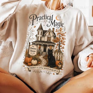 May include: A cream-colored crewneck sweatshirt featuring a graphic print of a Victorian-style house labeled The Owens House Est. 1998, a black cat, pumpkins, and autumn trees. The text reads Practical Magic and There's a little witch in all of us.