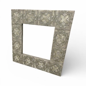 May include: A decorative silver-colored metal frame with a square opening. The frame is made of embossed tiles with a repeating floral pattern. The frame is angled, showcasing its ornate design and texture.