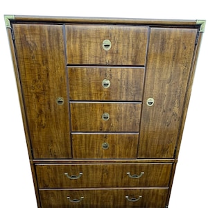 May include: A wooden armoire with a warm brown finish. The armoire features multiple drawers and cabinets with brass-toned hardware. The design includes a combination of drawers and doors, offering storage options. The wood grain is visible throughout the piece.