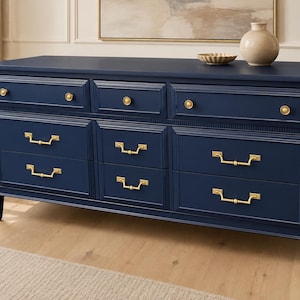 May include: A navy blue dresser featuring nine drawers with gold-toned hardware. The top row has three smaller drawers with round knobs, while the lower two rows contain six larger drawers with rectangular pull handles. The piece has tapered legs.