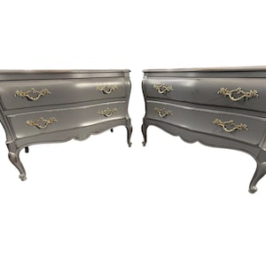 May include: Two matching gray dressers with curved fronts and two drawers each. The dressers have ornate gold-colored handles and legs. The tops of the dressers appear to be a darker color than the rest of the dresser.