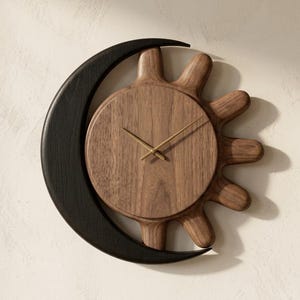 May include: A wall clock featuring a circular wooden sun face with a natural brown grain and thin gold hands, partially encircled by a smooth, curved black crescent moon shape. The design combines a dark wood sun and a black moon element.