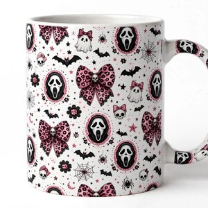 May include: A white ceramic mug featuring a repeating pattern of pink leopard print bows with small skulls, ghost figures with pink bows, black bats, spider webs, spiders, and framed screaming ghost masks with pink glittery borders.