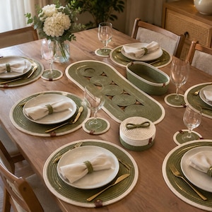May include: A dining table set featuring round olive green and white rope placemats, matching coasters, a long oval table runner, a small lidded storage basket, and a rectangular basket, all accented with small brown leather-like tabs.