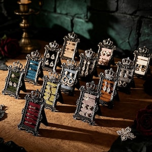 Peut inclure: Une collection de petits présentoirs à bijoux rectangulaires, chacun doté d'un cadre en métal noir orné surmonté d'un motif de couronne. Les présentoirs sont recouverts de velours de diverses couleurs, dont rouge, vert, bleu, jaune et beige, et accueillent des bagues et des boucles d'oreilles.