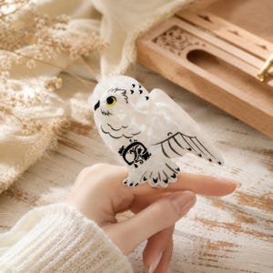 May include: A white acrylic hair claw clip shaped like a snowy owl with black etched wing and feather details, a yellow eye, and a decorative black floral scroll pattern on the chest, designed to hold hair securely with a spring-loaded mechanism.