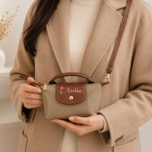 May include: A small beige handbag with a brown leather-textured flap, a top handle, and a detachable shoulder strap. The flap features a gold-tone snap button, an embroidered pink tulip, and the white cursive text Amlia printed on the front.