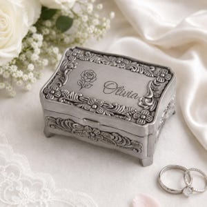 Könnte beinhalten: Eine rechteckige Aufbewahrungsbox aus silberfarbenem Metall mit vier kleinen Füßen. Der Deckel zeigt einen geprägten Blumenrand, eine gravierte Rosenillustration und den Namen Olivia in Schreibschrift. Zwei silberfarbene Metallringe.